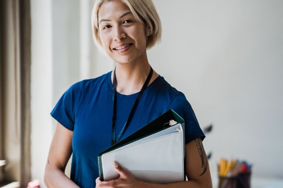 Enseignante souriante qui tient des classeurs contre elle, debout dans une salle de travail