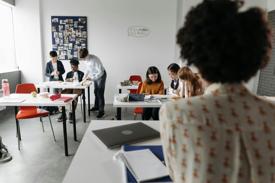 Enseignante de dos face à sa classe, où des élèves travaillent en petits groupes à plusieurs tables